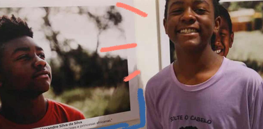 Foto de um menino negro sorrindo para a câmera. Atrás dele, há cartazes colocados na parede com fotos de pessoas negras.
