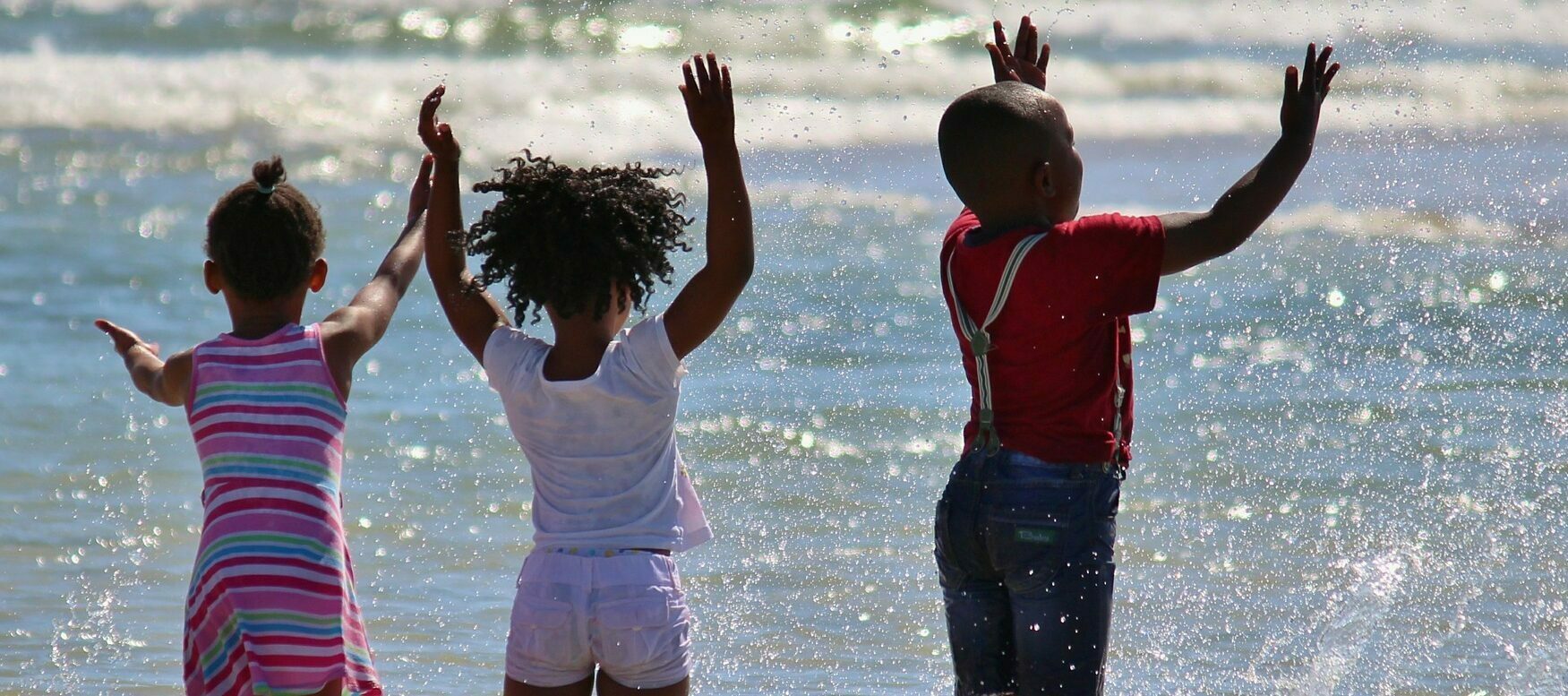 Três crianças negras brincam no mar. Elas jogam a água do mar para cima e esticam os braços.
