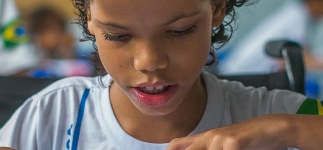 Foto de uma criança em uma carteira escolar. Ela olha para baixo e segura um lápis.