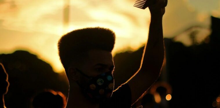 Foto de um jovem negro em um protesto. Ele usa máscara de proteção, segura um cartaz e está com uma das mãos para cima.
