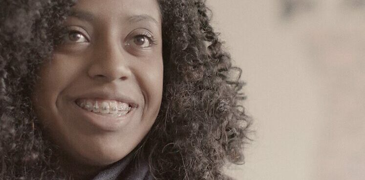 Foto de uma menina negra de cabelos cacheados. Ela sorri e usa aparelho nos dentes.
